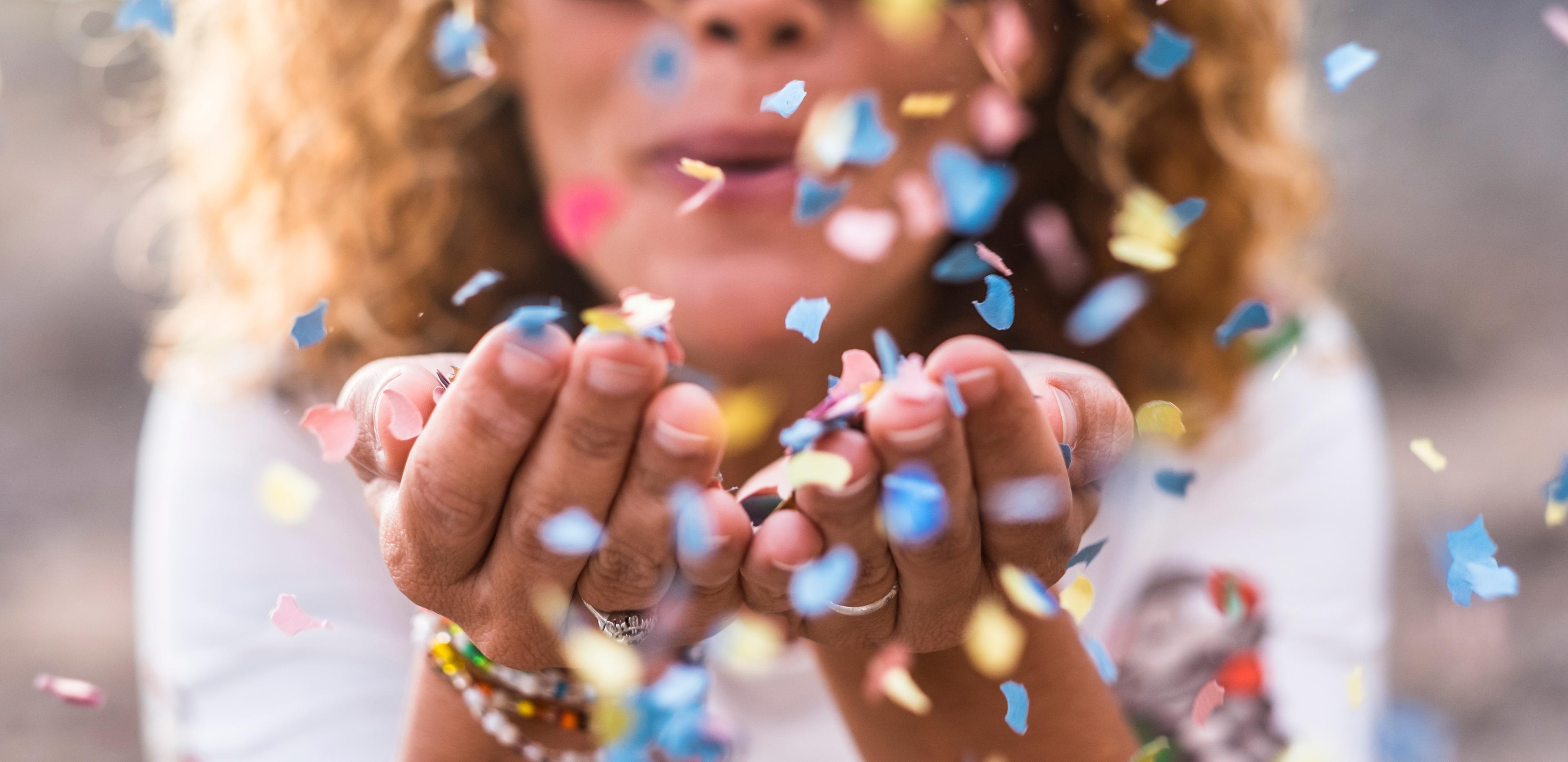 Een persoon blaast kleurrijke confetti uit haar handen. De focus ligt op de handen en de confetti, terwijl het gezicht van de persoon enigszins wazig is op de achtergrond.  